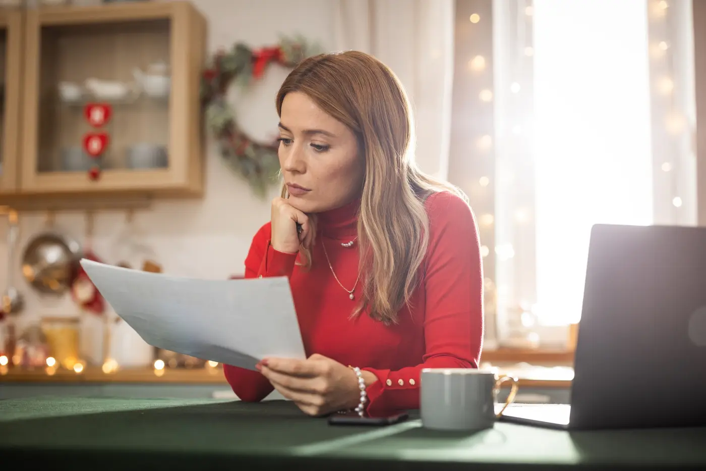 A woman wearing a red sweater sits in a holiday-decorated kitchen, intently examining a document in her hands.