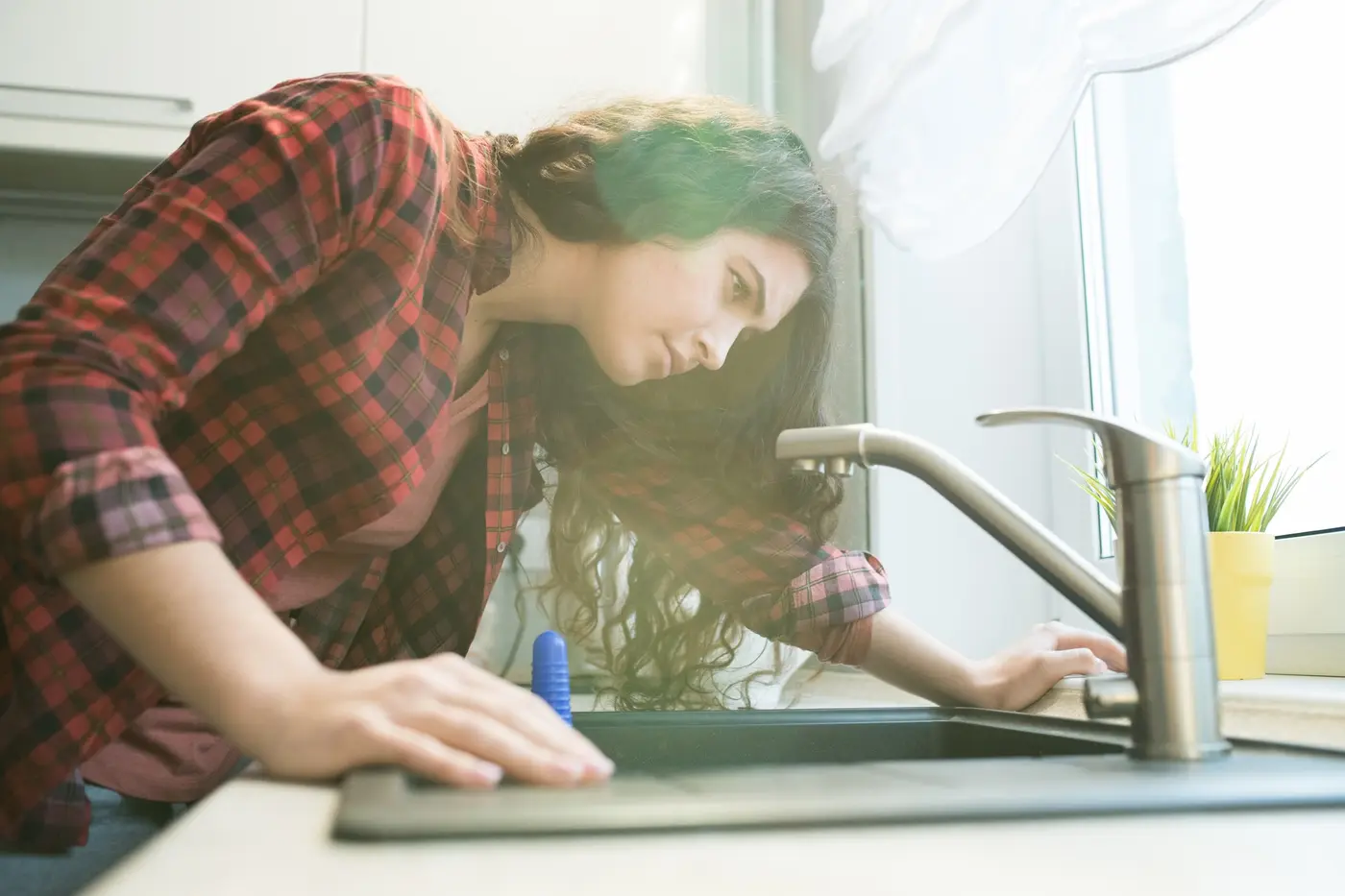 Woman in checkered shirt checking faucet while having problem with dripping faucet in kitchen.