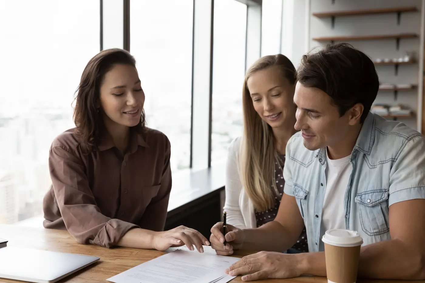 Happy young couple signs a document to finalize their agreement with a female property manager in the office.